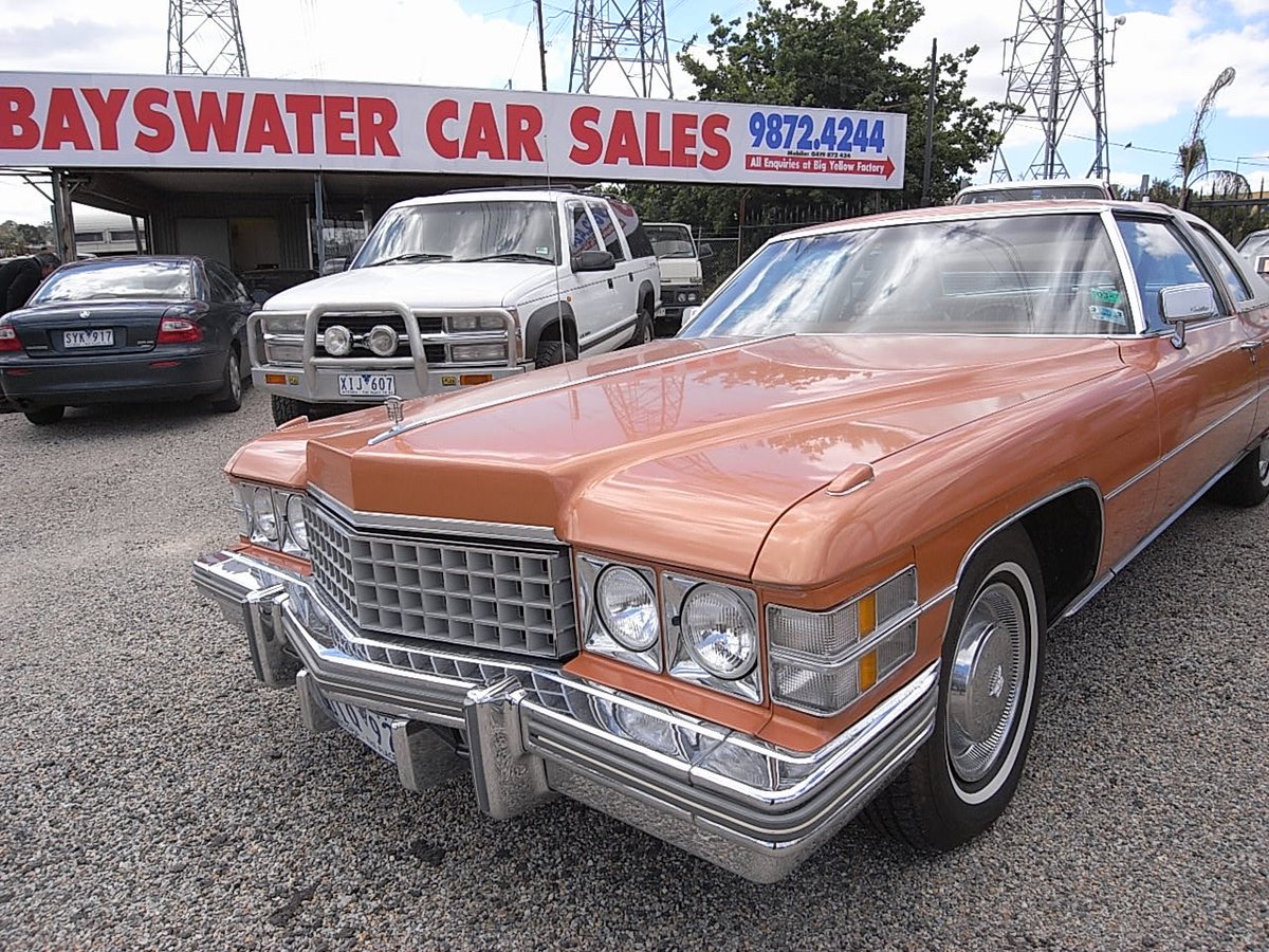 A classic Cadillac coupe parked outside, underneath the large Bayswater Car Sales roadside sign